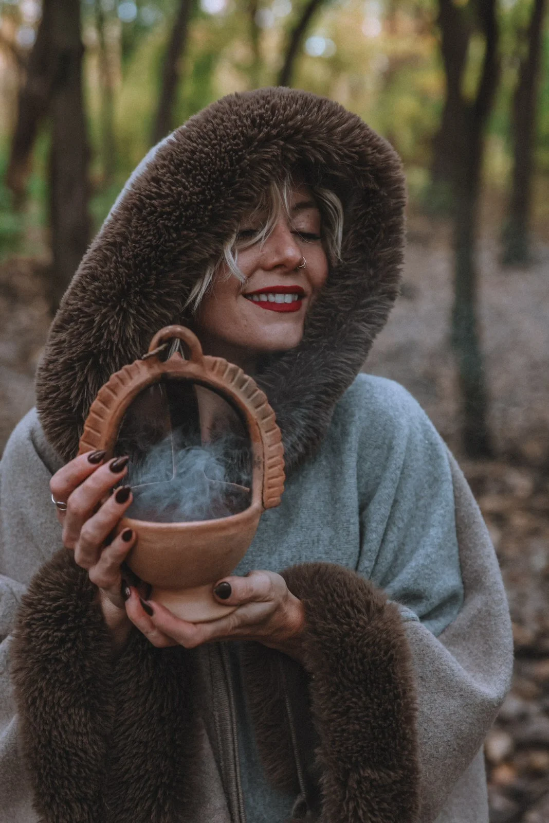 Christina Cox smiling, holding a smoking clay bowl in the forest