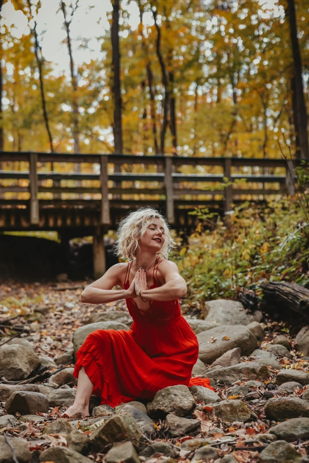 Christina in a red dress with hands in prayer by a creek in autumn