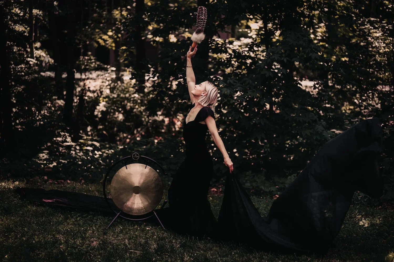 Christina raising a smoking smudge bowl beside a gong in the woods at dusk